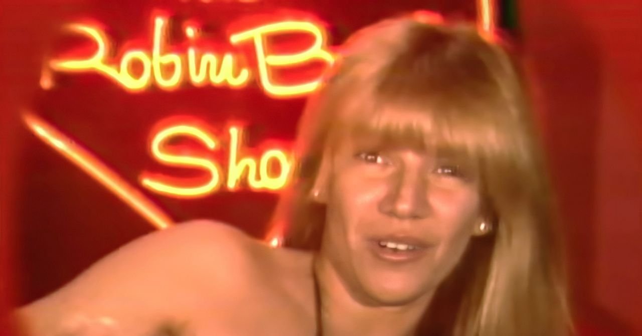 Robin Byrd, the iconic public access TV star, smiling in a black mesh bikini on her show's heart-shaped set.