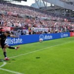 A soccer ball curving through the air towards a goal, with a blurred background of a stadium.