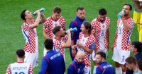 Football players taking a hydration break during a World Cup match, with advertising hoardings visible.