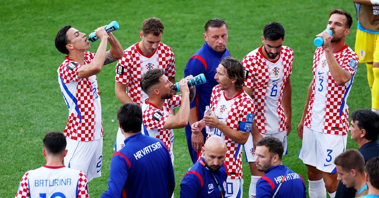 Football players taking a hydration break during a World Cup match, with advertising hoardings visible.