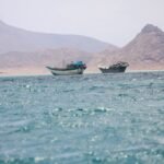 قوارب صغيرة تطفو بالقرب من ساحل باب المندب في اليمن، مع ناقلات نفط في الأفق، تعكس التوترات الجيوسياسية التي تؤثر على أسعار النفط العالمية.