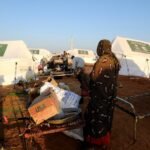 امرأة نازحة تقف أمام صندوق كرتوني فارغ في مخيم للنازحين بمدينة الأبيض، السودان، في مشهد يعكس الأزمة الإنسانية المتفاقمة.
