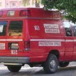 صورة لدراجة نارية سقطت في ساقية للري في منطقة ريفية، مع وجود سيارات إسعاف وسلطات في الخلفية.