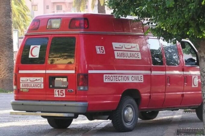 صورة لدراجة نارية سقطت في ساقية للري في منطقة ريفية، مع وجود سيارات إسعاف وسلطات في الخلفية.