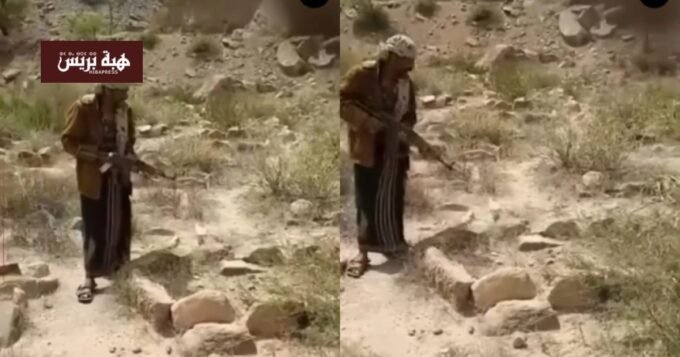شاب يمني يحمل بندقية ويطلق النار على قبر في منطقة جبلية، تعبيرًا عن غضبه من نزاع على الميراث.