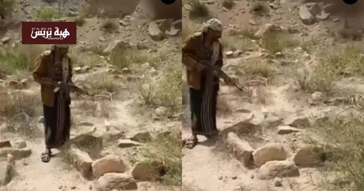 شاب يمني يحمل بندقية ويطلق النار على قبر في منطقة جبلية، تعبيرًا عن غضبه من نزاع على الميراث.