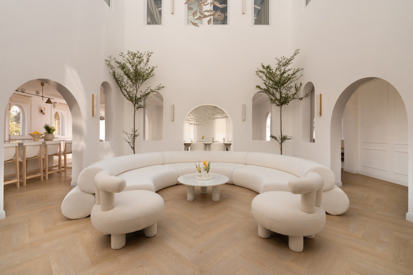A bright, open central courtyard of The White Arcade luxury home in Dubai, featuring layered white arches, a glass roof, mature trees, and a curved bespoke sofa.