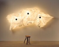 Erwan Bouroullec's Maap wall lamp, showcasing a crumpled Tyvek diffuser secured by magnetic dots on a steel framework, casting a soft, ambient glow on a wall.