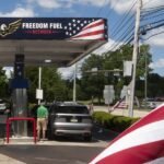 A Freedom Fuel Network gas station pump displaying low prices, with a blurred image of Donald Trump in the background.