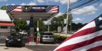 A Freedom Fuel Network gas station pump displaying low prices, with a blurred image of Donald Trump in the background.