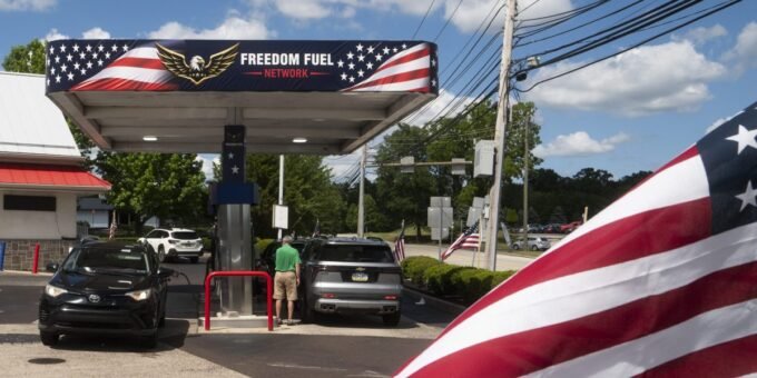 A Freedom Fuel Network gas station pump displaying low prices, with a blurred image of Donald Trump in the background.