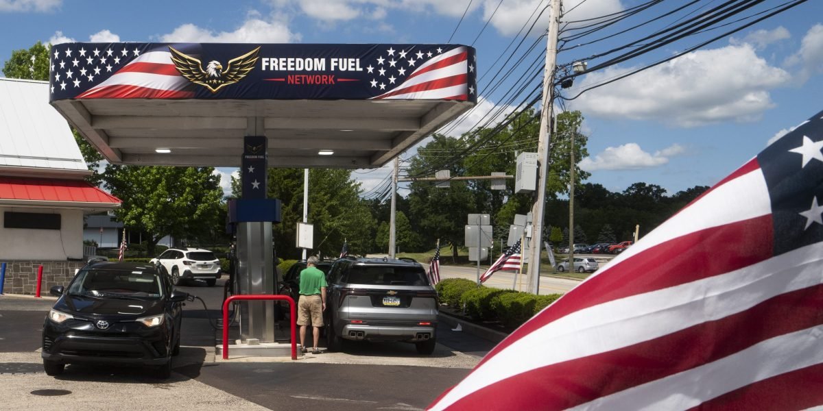 A Freedom Fuel Network gas station pump displaying low prices, with a blurred image of Donald Trump in the background.