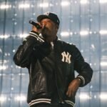 Jay-Z performing on a minimalist stage at Yankee Stadium, backlit by a large screen displaying early New York images.
