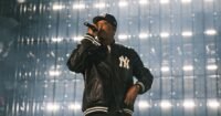 Jay-Z performing on a minimalist stage at Yankee Stadium, backlit by a large screen displaying early New York images.