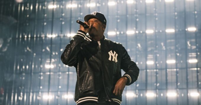Jay-Z performing on a minimalist stage at Yankee Stadium, backlit by a large screen displaying early New York images.