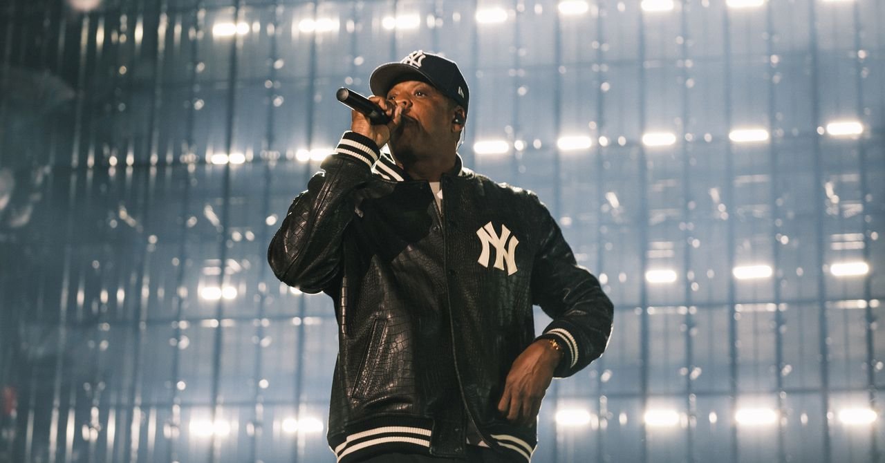 Jay-Z performing on a minimalist stage at Yankee Stadium, backlit by a large screen displaying early New York images.
