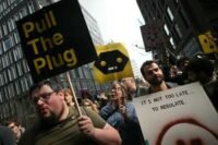 A diverse group of people gathered in Washington Square Park, some holding up 'dumbphones' or signs, listening to a speaker, symbolizing a movement against pervasive digital technology.