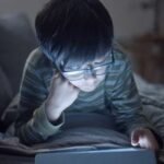 More Than Meets the Eye: The Systemic Health Risks of Screen Time 8 Person looking at a smartphone screen at night, with blue light emitting, illustrating the impact of screens on health.
