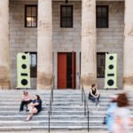 Seven-foot-tall sculptural speakers crafted from Baltic birch plywood and MDF mounted on the historic facade of the Village of Industry & Art building in Philadelphia, broadcasting music.