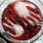 A vibrant red Halloween punch bowl featuring a frozen hand ice cube, garnished with dark frozen cherries, set against a spooky party backdrop.