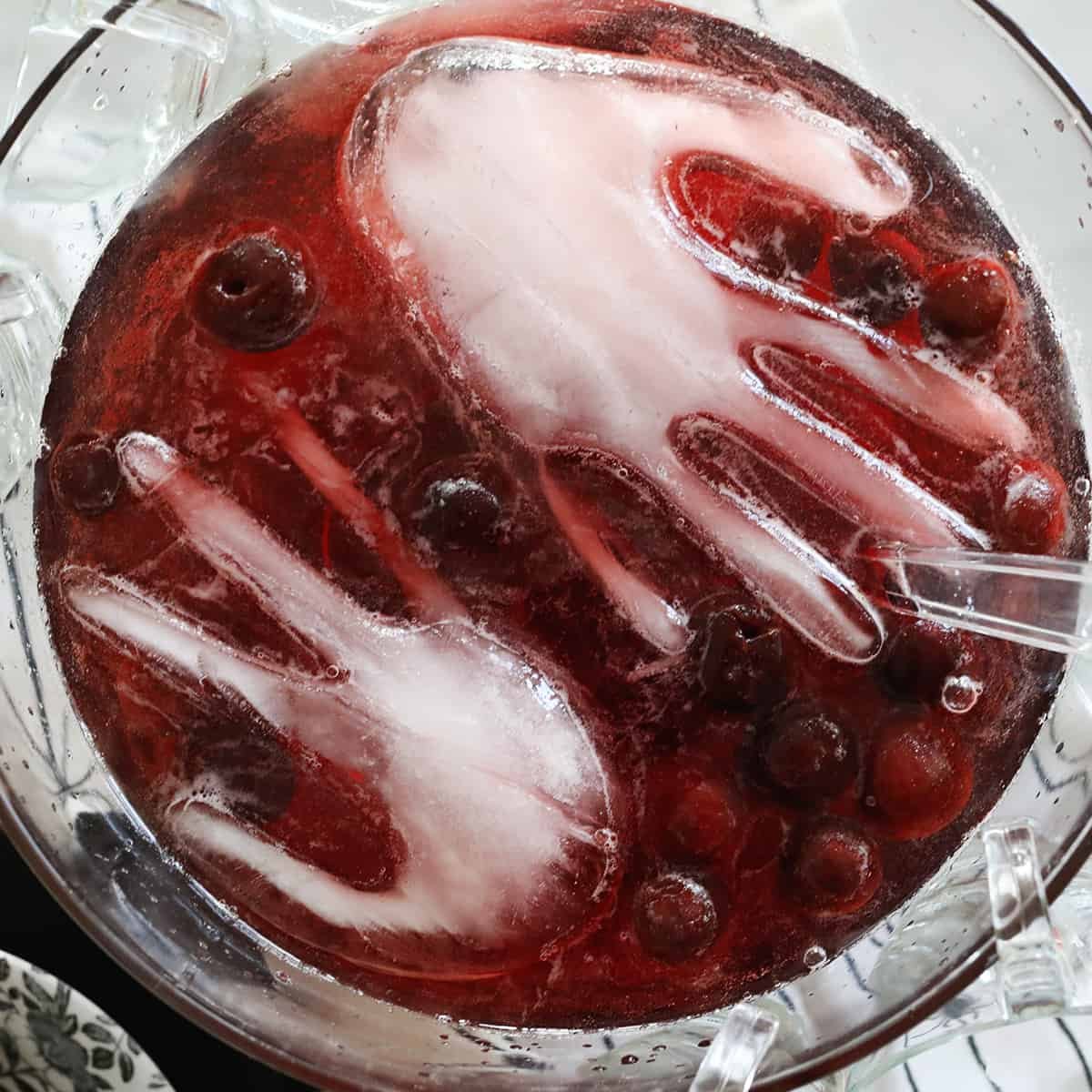 A vibrant red Halloween punch bowl featuring a frozen hand ice cube, garnished with dark frozen cherries, set against a spooky party backdrop.
