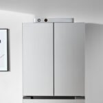 Jackery FridgeGuard, a sleek silver ultra-slim smart battery, standing next to a stainless steel refrigerator in a modern kitchen.
