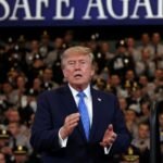 Two youths on the shoreline of Bandar Abbas, Iran, with ships anchored in the Strait of Hormuz, and U.S. President Donald Trump delivering remarks in Garden City, New York.