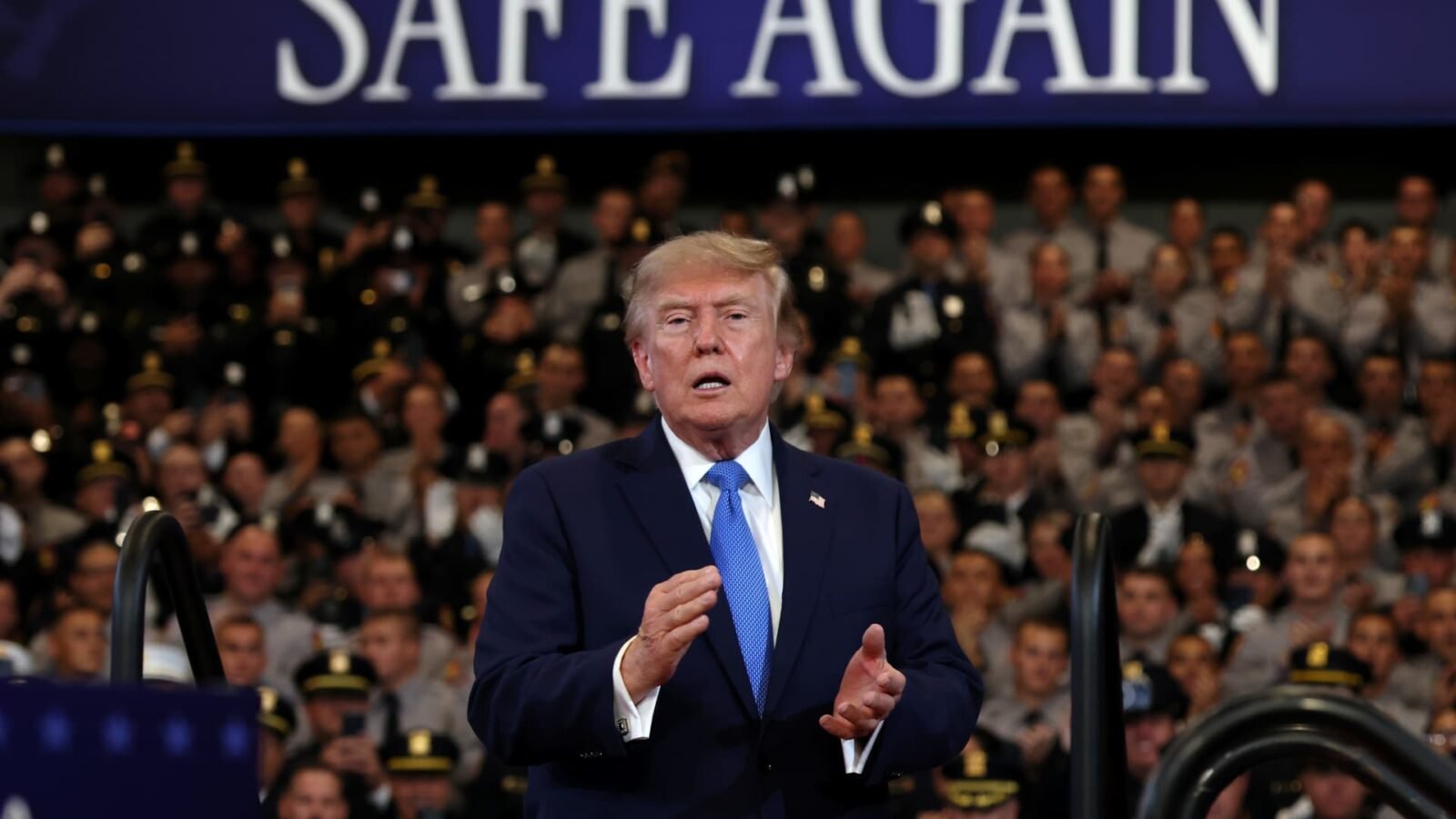 Two youths on the shoreline of Bandar Abbas, Iran, with ships anchored in the Strait of Hormuz, and U.S. President Donald Trump delivering remarks in Garden City, New York.