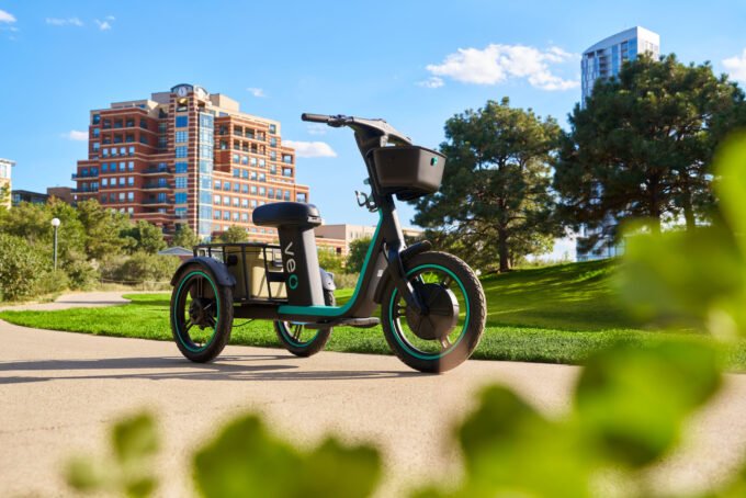 Veo's Rover electric trike, featuring three wheels, a cushioned seat, and baskets, in an urban setting.