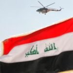 صورة جوية للعلم العراقي يرفرف فوق قاعدة عسكرية في بغداد، مع مروحية تحلق في السماء.