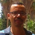 سائق أجرة مغربي ذو شعر أبيض يفكر بعمق بينما امرأة مسنة تنزل من سيارته في مدينة مغربية.