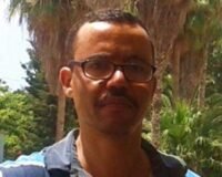 سائق أجرة مغربي ذو شعر أبيض يفكر بعمق بينما امرأة مسنة تنزل من سيارته في مدينة مغربية.
