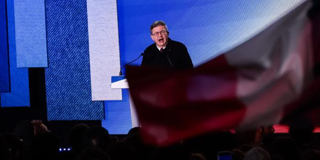 Jean-Luc Mélenchon s'adressant à la foule lors de la Fête de l'Humanité, avec des drapeaux de La France Insoumise et de la NUPES en arrière-plan, symbolisant son appel à l'unité de la gauche pour 2027.