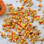 A close-up of golden-brown roasted pumpkin seeds on a baking sheet, some mixed with colorful candy corn.