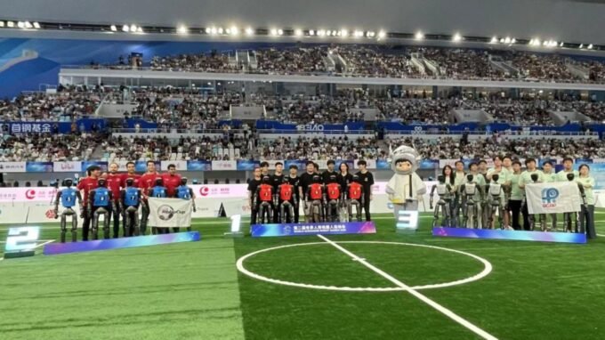 Two humanoid robots playing football on a green pitch, with one robot kicking the ball and another in pursuit, demonstrating advanced robotics and AI.