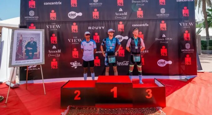Agadir Vibre au Rythme de l'IRONMAN 70.3 : Aziz Haddaj sur le Podium, un Éclat International 9 Vue aérienne de triathlètes nageant dans la baie d'Agadir lors de l'IRONMAN 70.3, avec la ville et les montagnes en arrière-plan, symbolisant le défi sportif et le cadre marocain.