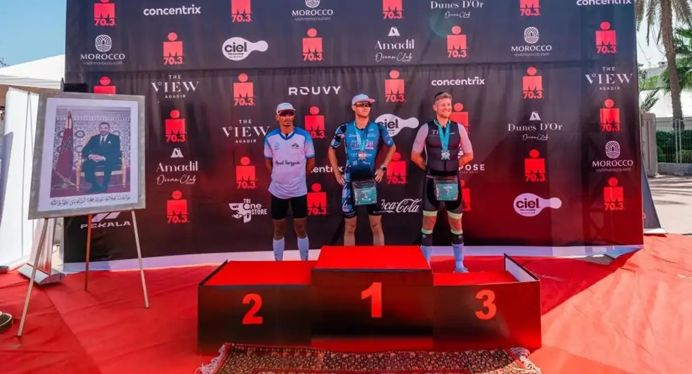 Vue aérienne de triathlètes nageant dans la baie d'Agadir lors de l'IRONMAN 70.3, avec la ville et les montagnes en arrière-plan, symbolisant le défi sportif et le cadre marocain.