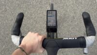 Un cycliste utilisant le compteur GPS Coros Dura 2 Solar sur son vélo, avec un paysage en arrière-plan. L'écran du compteur affiche des données de performance.