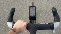 Un cycliste utilisant le compteur GPS Coros Dura 2 Solar sur son vélo, avec un paysage en arrière-plan. L'écran du compteur affiche des données de performance.