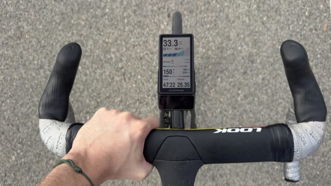 Un cycliste utilisant le compteur GPS Coros Dura 2 Solar sur son vélo, avec un paysage en arrière-plan. L'écran du compteur affiche des données de performance.