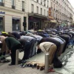 مصلون يؤدون صلاة الجمعة في شارع مغلق بجوار مسجد، مع حركة مرور متوقفة.