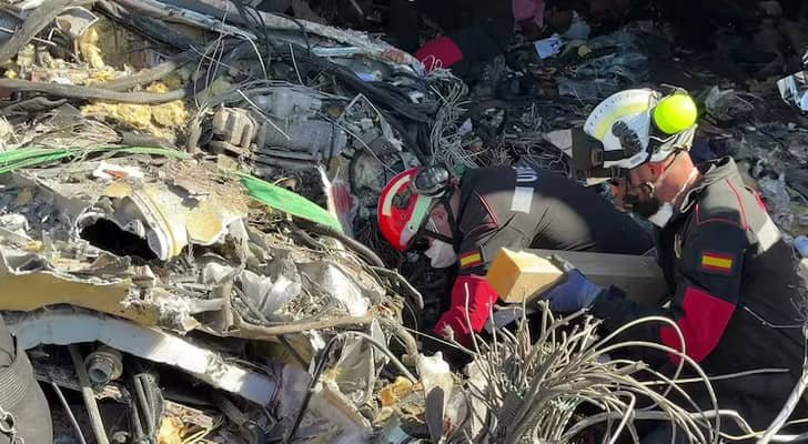 صورة لحطام قطار بعد تصادم مأساوي في إسبانيا، تظهر عربات متضررة وسيارات إسعاف في الخلفية.