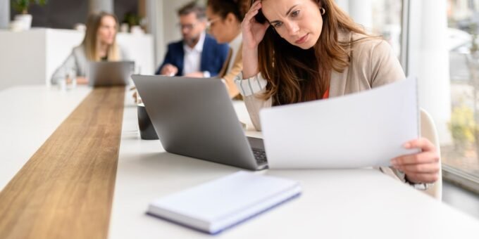 An overwhelmed employee juggling multiple tasks at a desk, symbolizing the 'two-job job' phenomenon.