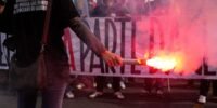 Riot police facing off against young protesters in a European city, with luxury stores boarded up in the background.