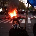 A protestor wearing a gas mask holds a French flag in front of a burning barricade during a demonstration in Paris against the high cost of living.