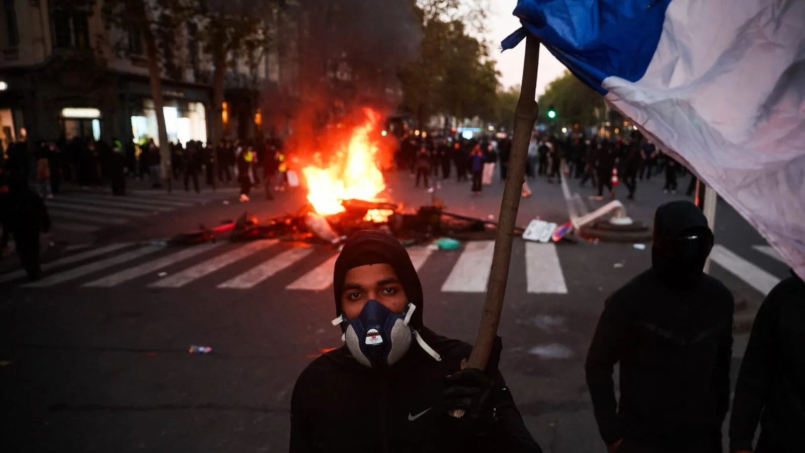 A protestor wearing a gas mask holds a French flag in front of a burning barricade during a demonstration in Paris against the high cost of living.