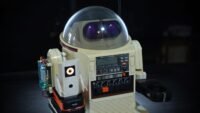 A vintage Tomy Omnibot robot with a modern 3D-printed AI companion, Ring-O, on its arm, looking at a laptop.