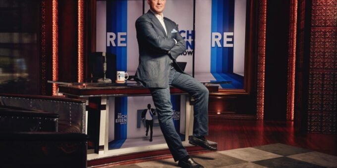 Rich Eisen smiling in a broadcast studio, surrounded by sports media equipment