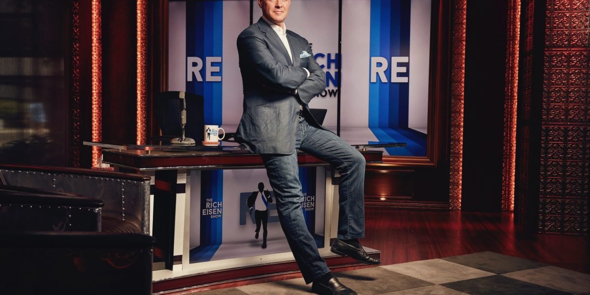 Rich Eisen smiling in a broadcast studio, surrounded by sports media equipment