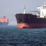 Oil tanker navigating the Strait of Hormuz with a naval vessel in the background, symbolizing the precarious security situation.